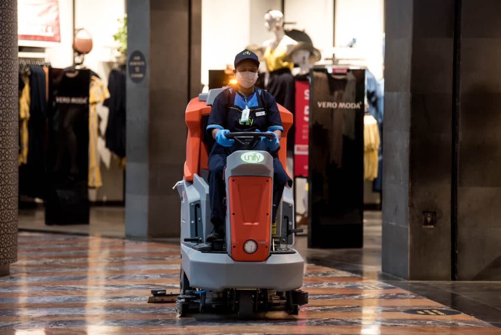 A diverse scene unfolds in a bustling shopping mall, featuring a person on a robot, workers using cleaning machines, and fashion displays.