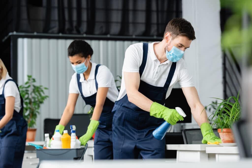 Group of individuals cleaning office, woman in overalls with mask and gloves, man in white shirt and surgical mask, person in green gloves with blue spray bottle.
